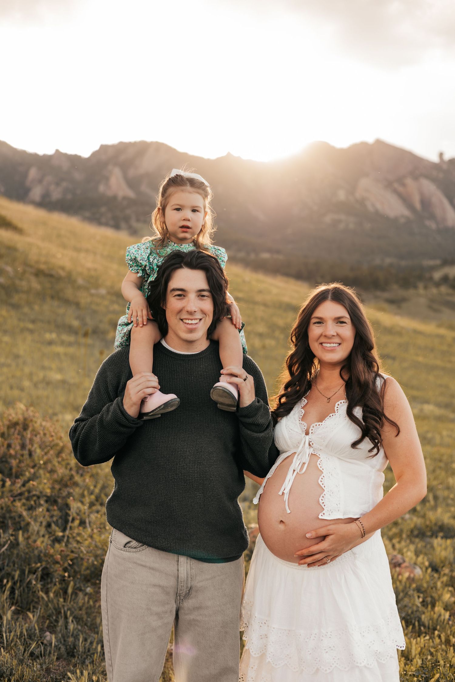Happy  family poses outdoors with toddler on dad's shoulders, pregnant mom in white dress, mountains | Boulder Family Photographer