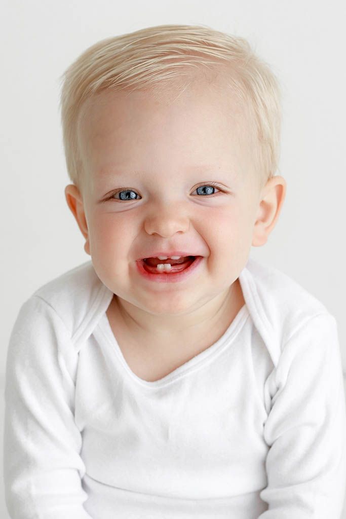 Smiling baby with blue eyes and blonde hair wearing a white onesie, showing first teeth against white background.