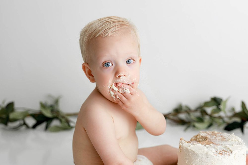 Blonde baby boy eating smash cake with frosting on face during first birthday photo session by Riverside cake smash photographer.