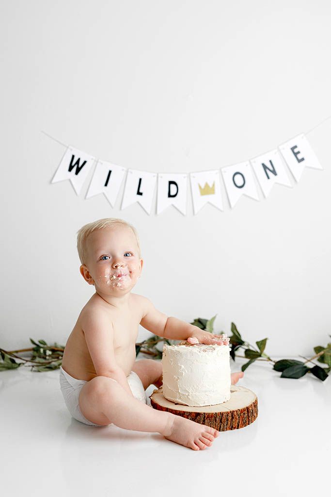 Baby's first birthday cake smash photo with Wild One banner, white frosted cake on wood slice, white background.