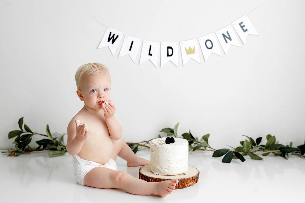 Baby in diaper sits by smash cake under Wild One banner with greenery for first birthday photo session by Riverside cake smash photographer.