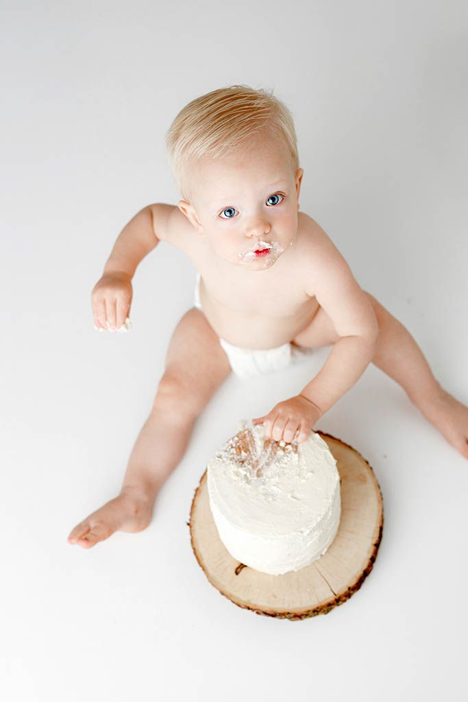 Blonde baby in diaper touching white frosted cake on wood slice during first birthday cake smash photo session.