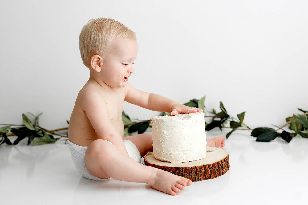 Baby in diaper touching white frosted cake on wood slice, surrounded by greenery, during first birthday cake smash photo session.