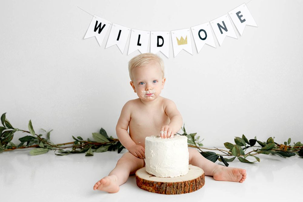 Baby boy sits with a white cake on a wood slice under a Wild One banner for a first birthday cake smash photo.