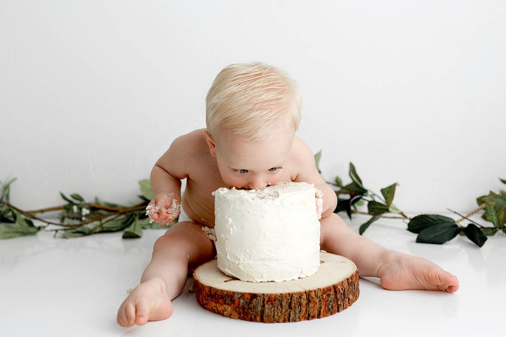 A baby enjoys their first birthday cake smash on a wooden slice against a white background with greenery.