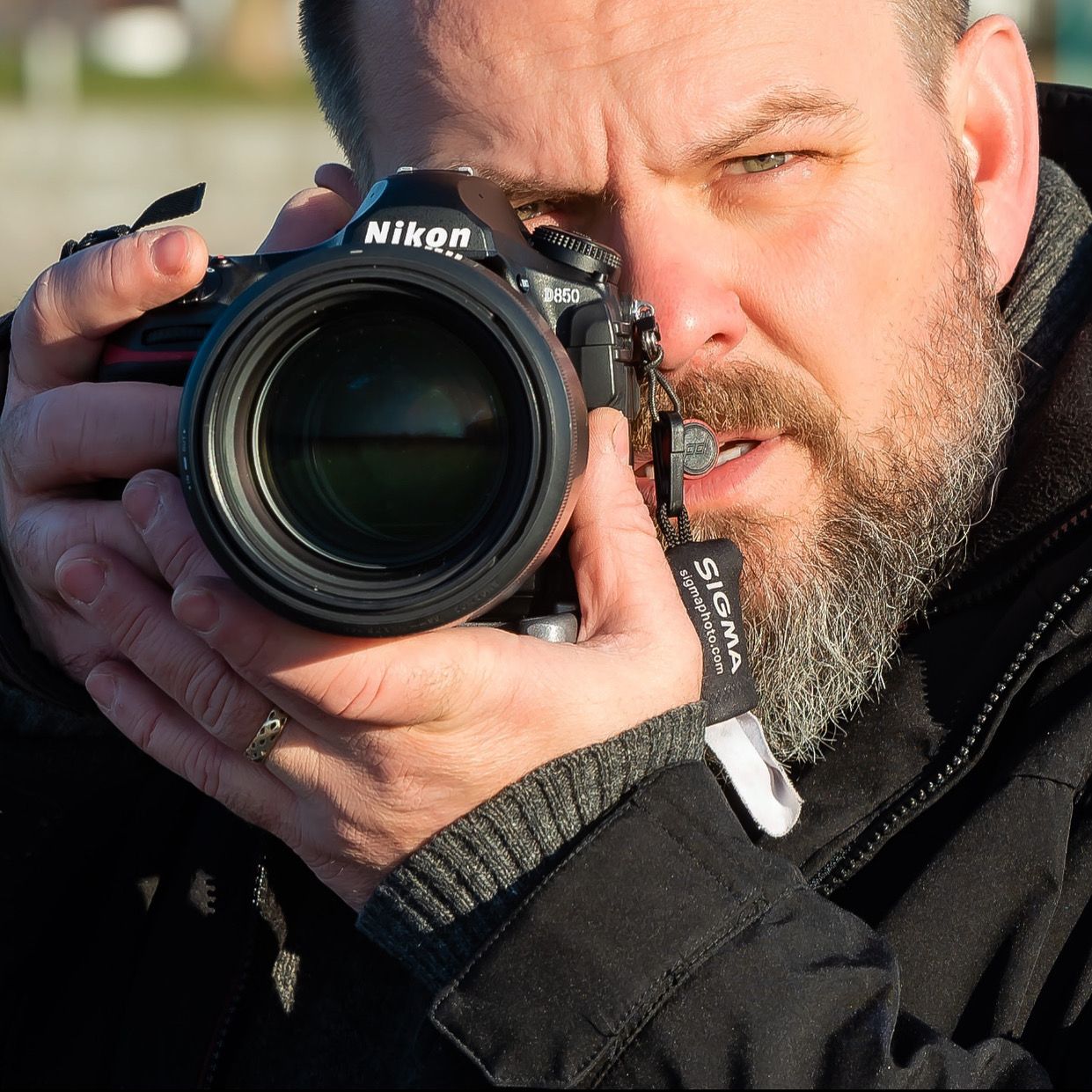 Bearded man holding a Nikon camera with Sigma lens up to his eye, photographing outdoors in natural light.