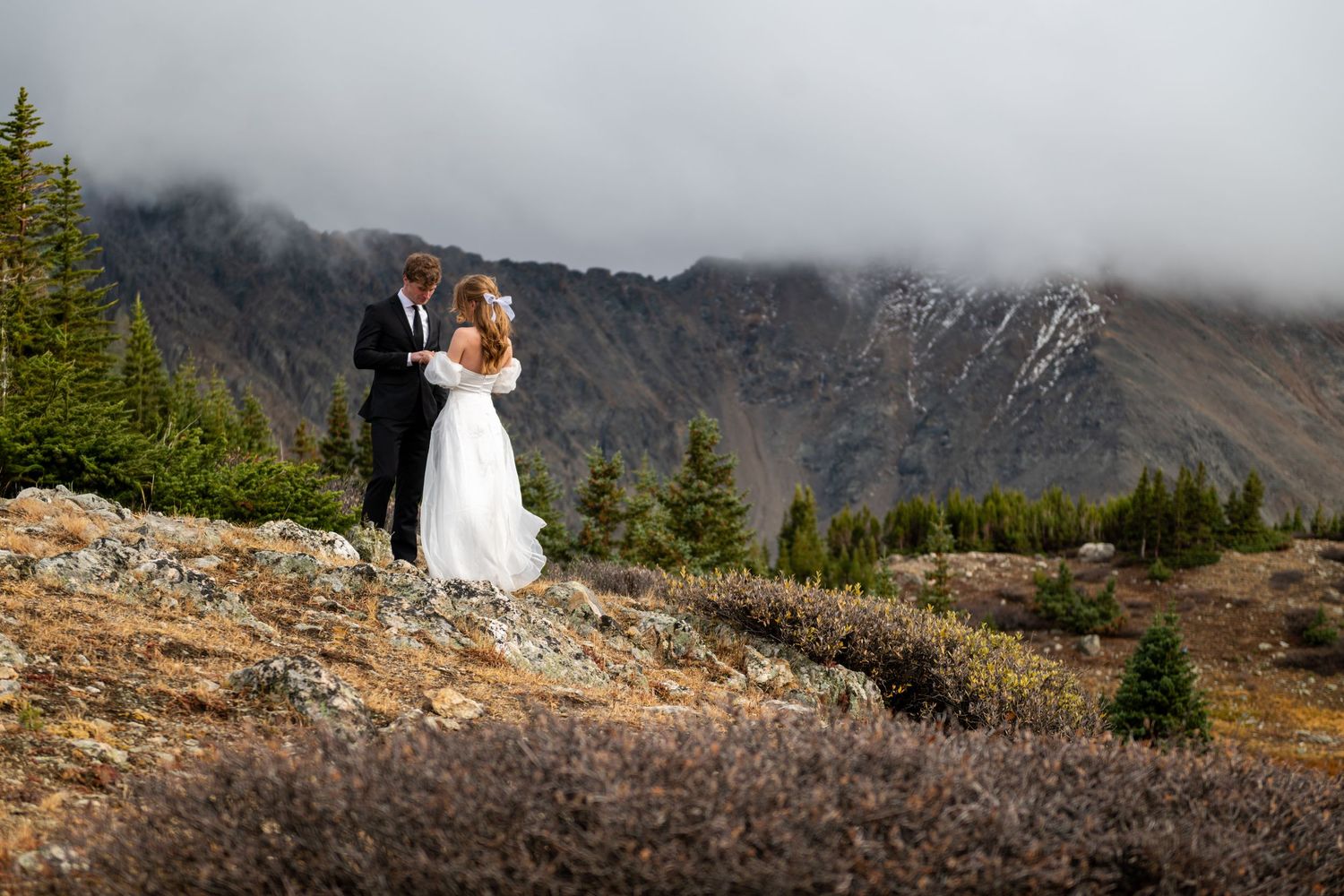 Bride and groom embrace on rocky mountain overlook with misty peaks and evergreen trees in background.