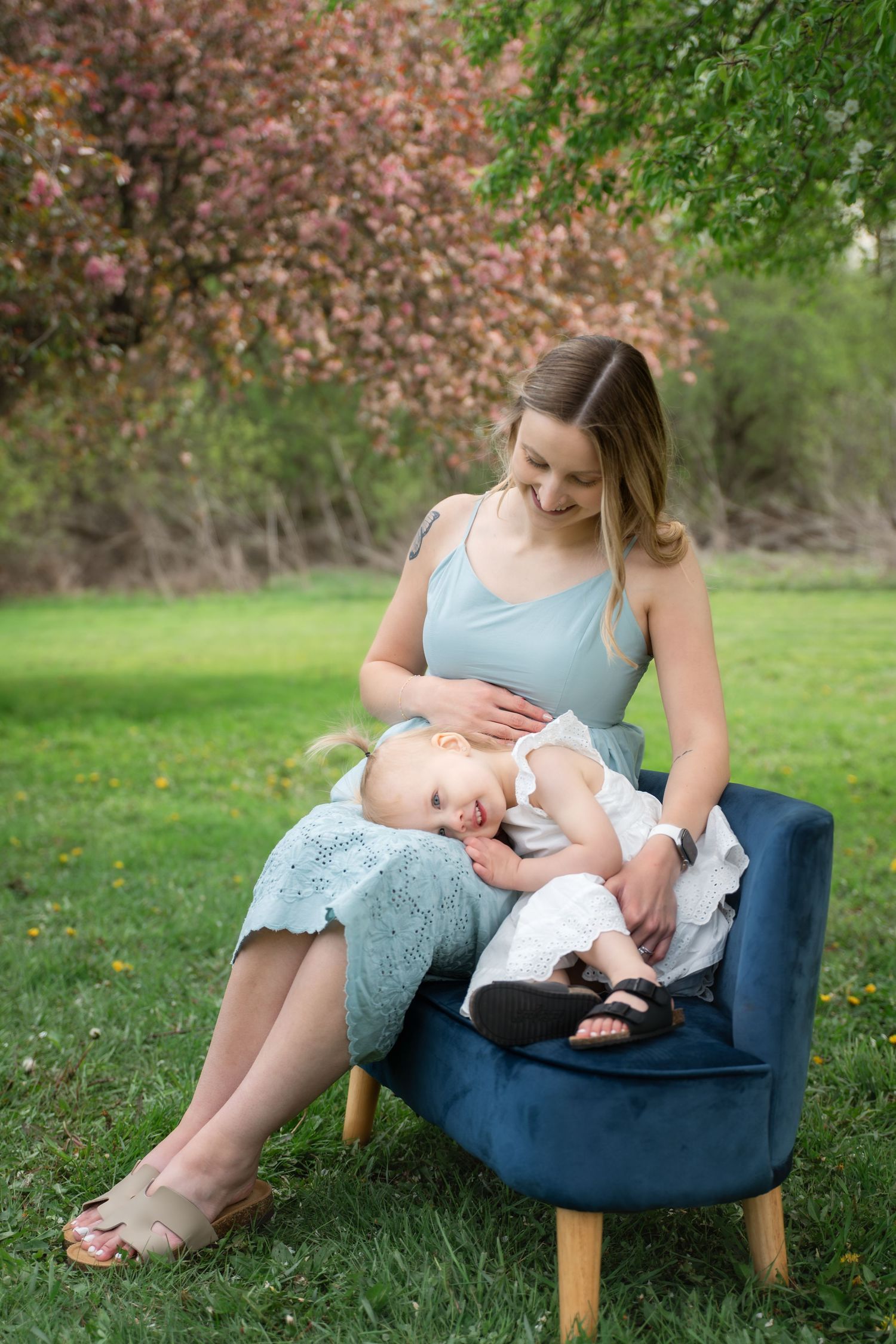 A young mother in a light blue dress sits on a navy velvet chair outdoors, lovingly holding her baby girl on her lap.