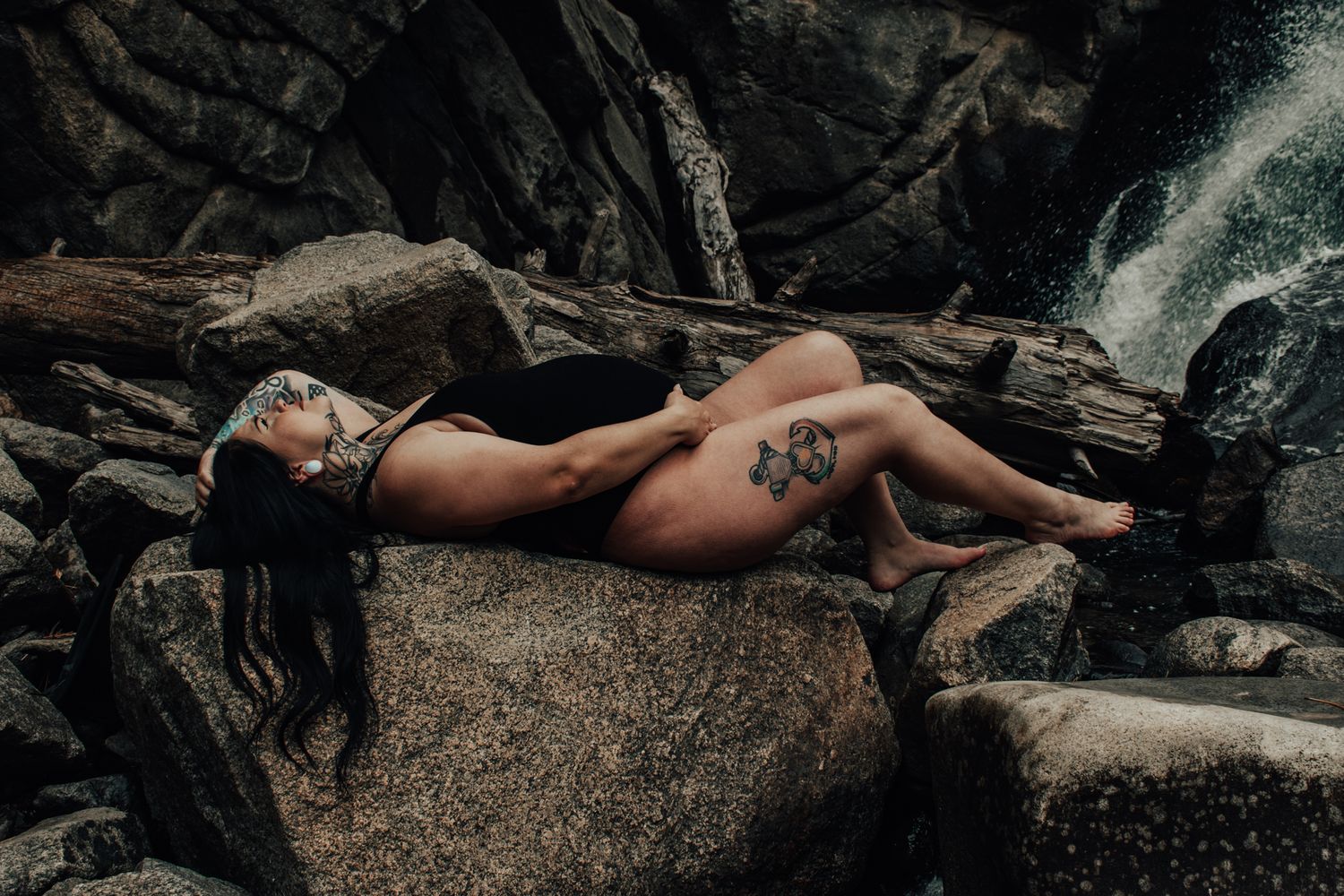 Woman with tattoos reclines on rocky terrain near a waterfall in a dramatic outdoor nature setting.