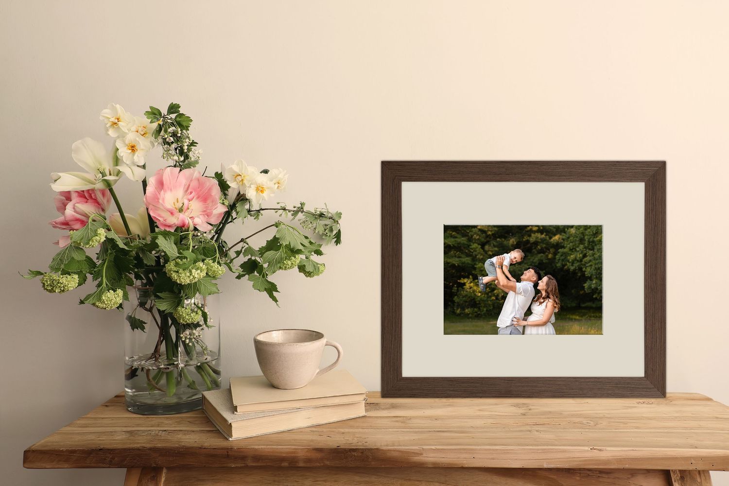 Framed family photo on a wooden table beside a vase of pink flowers and a beige coffee cup.