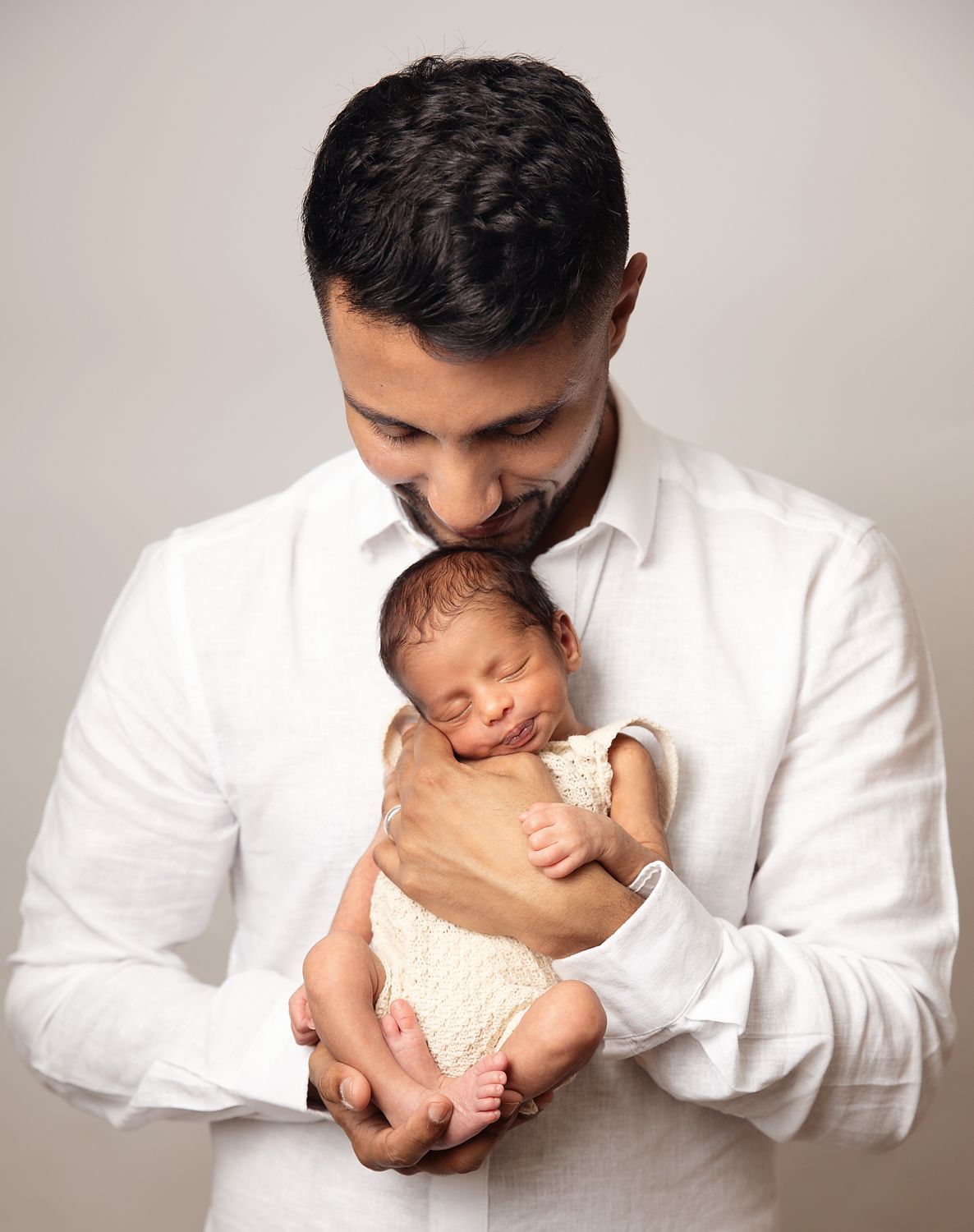 A proud father in a white shirt tenderly holds his sleeping newborn baby, both dressed in white. During a newborn session in Orpington