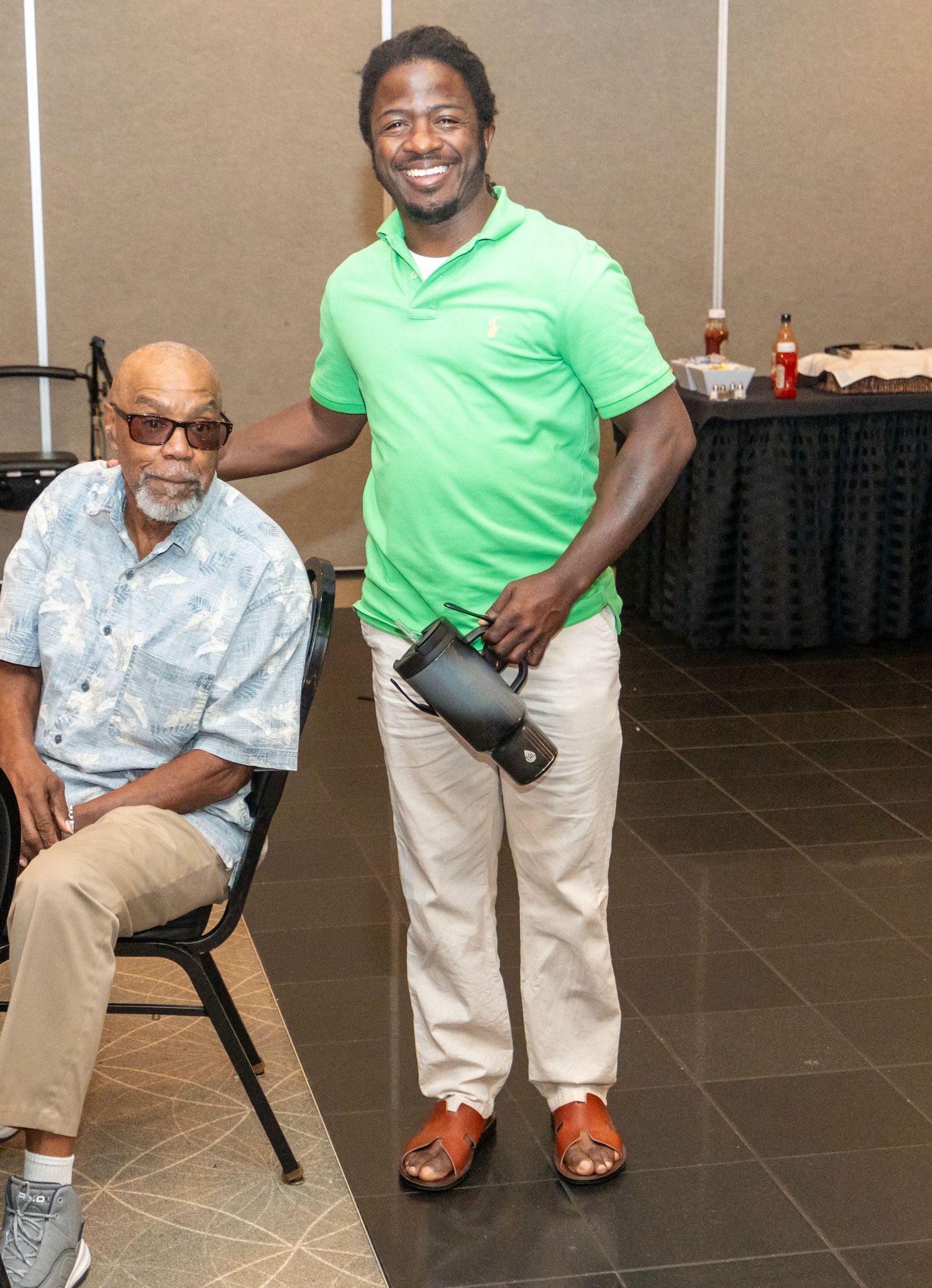 Two men pose together at an indoor event, one seated in a blue shirt and one standing in a green polo holding a bag.