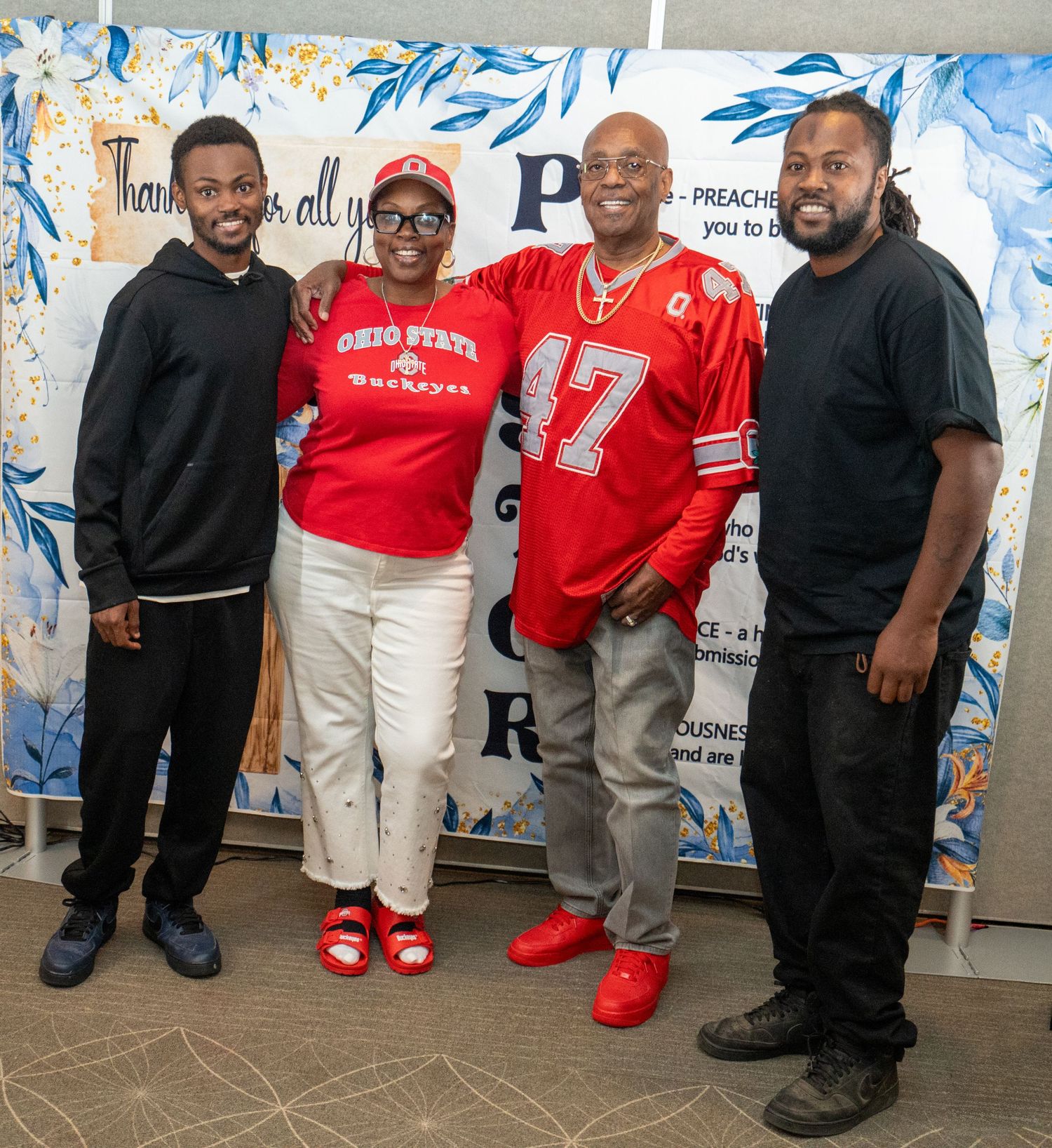 Four people posing together in Ohio State Buckeyes red apparel in front of a decorative blue floral backdrop.