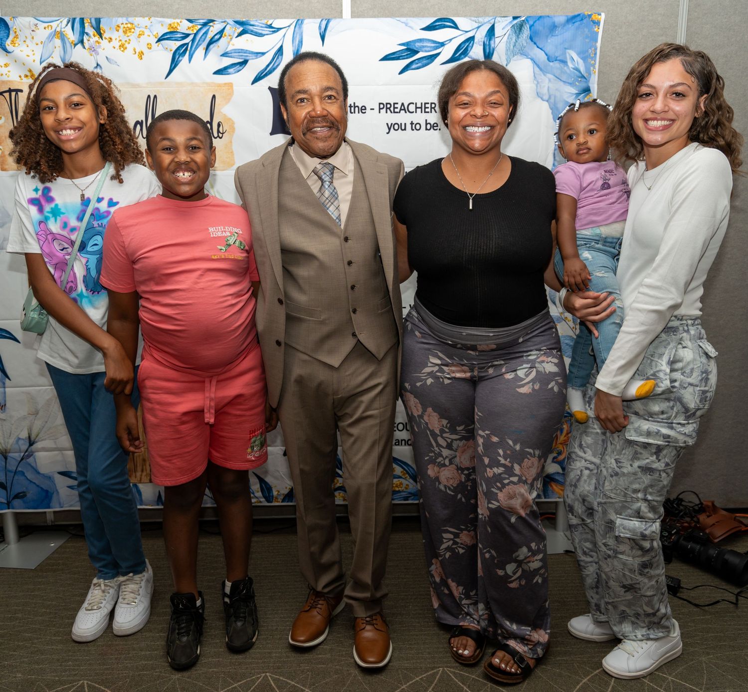 Group of six people posing together indoors at an event, smiling for a photo with a decorative banner behind them.