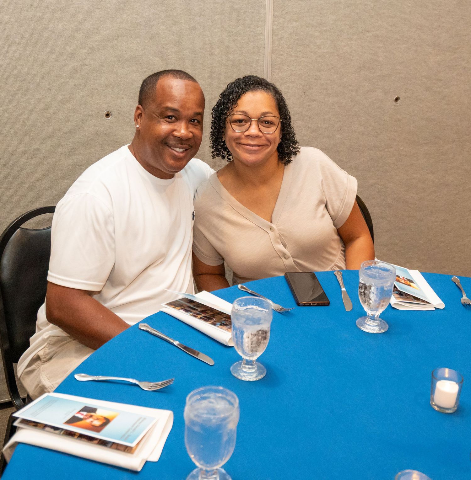 Smiling couple seated at a blue tablecloth dining table with water glasses and menus at a formal event.