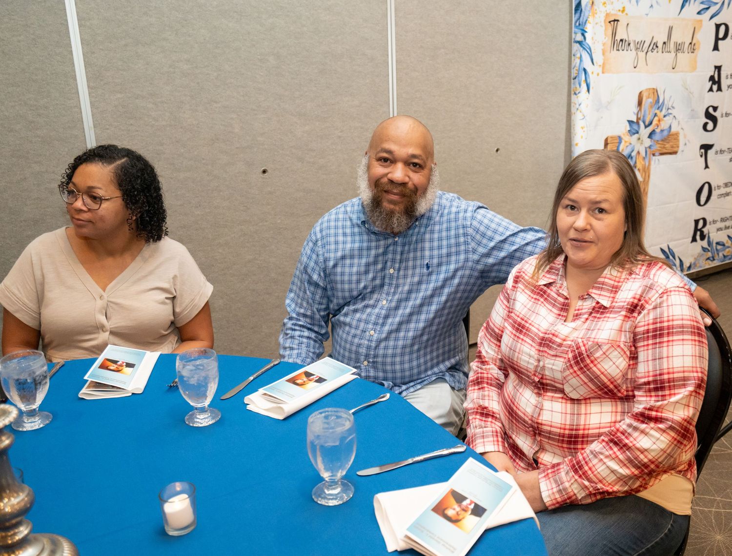 Three people smile at a blue table during a pastor appreciation event in a banquet hall.