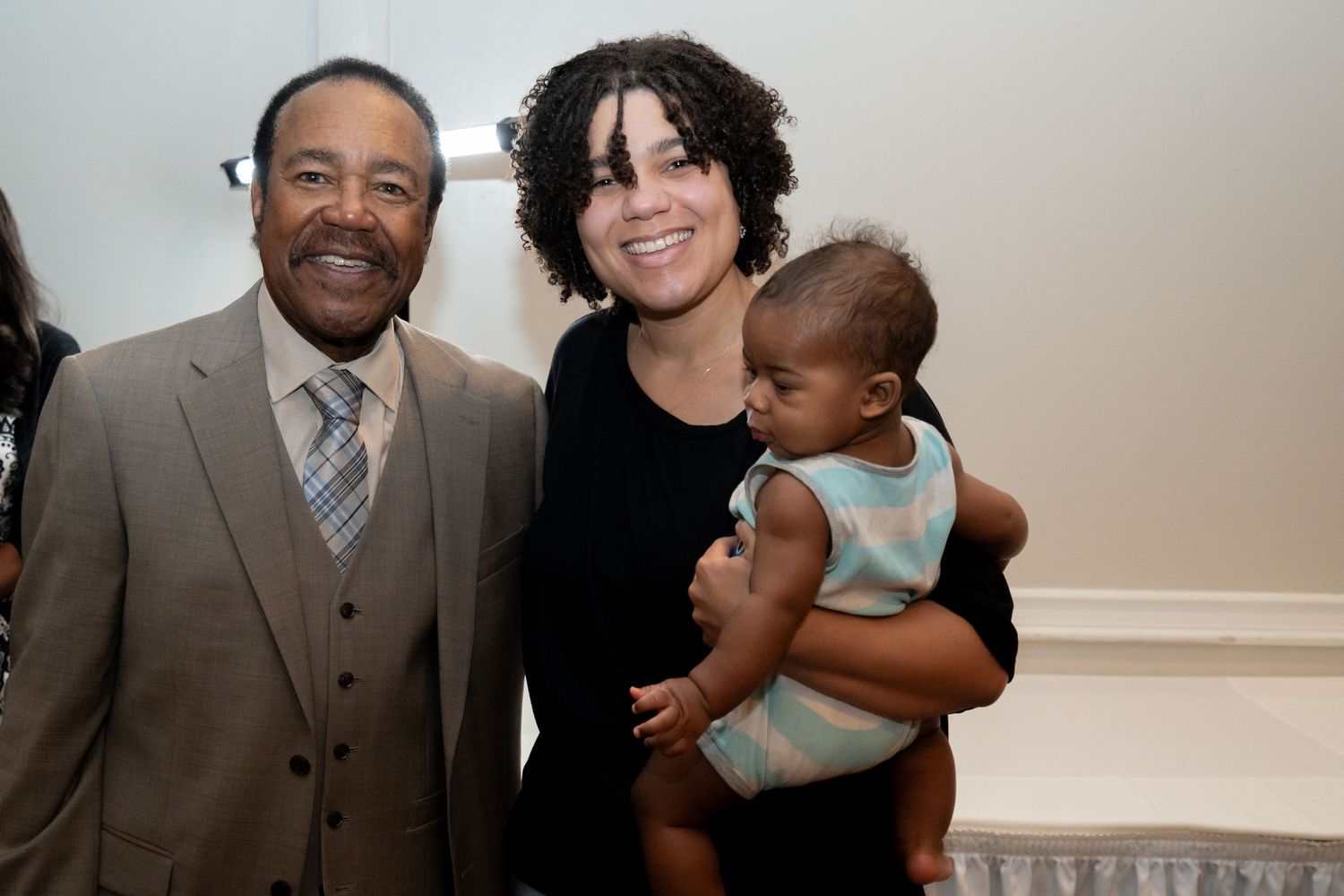 An elderly man and a young woman holding a baby pose together smiling at an indoor event.