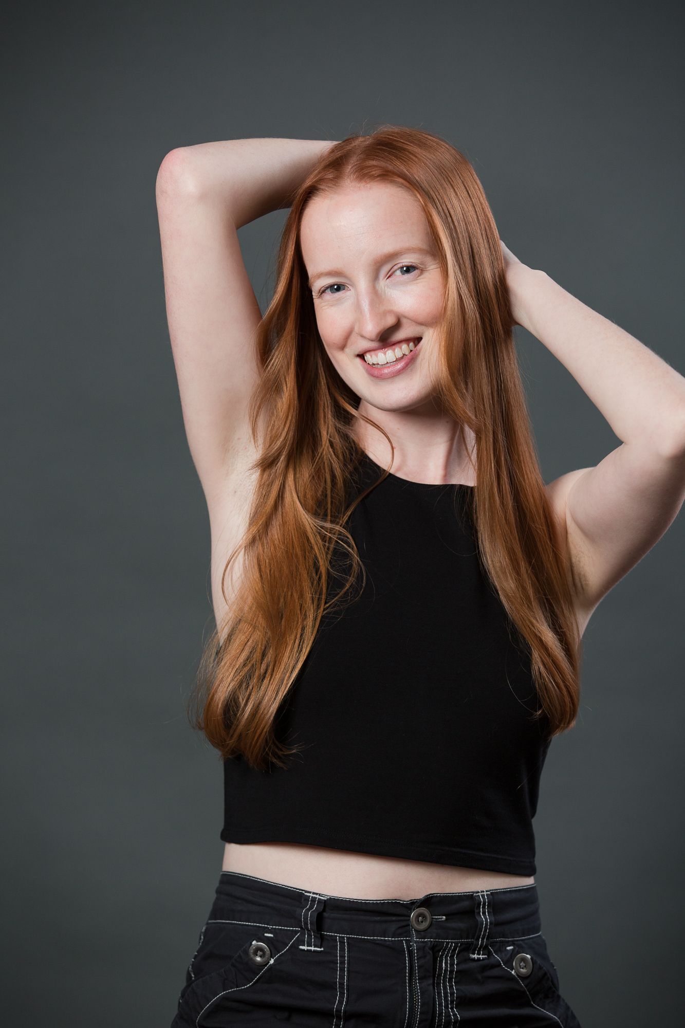Smiling young woman with long red hair wearing a black crop top and jeans, posing with hands behind head.