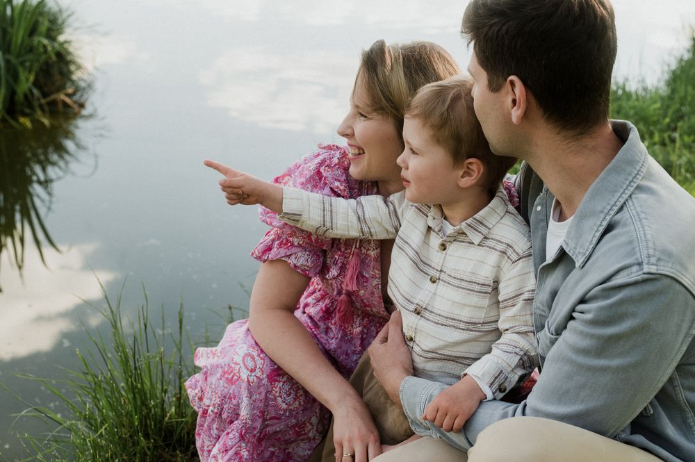 Family of three sitting by a pond, mother pointing at water while father kisses young child's head.