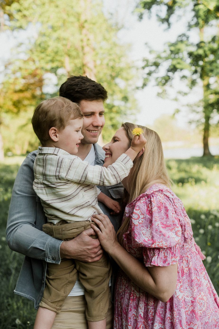 Happy family portrait outdoors, parents smiling at toddler son in green park setting, woman in pink floral dress.