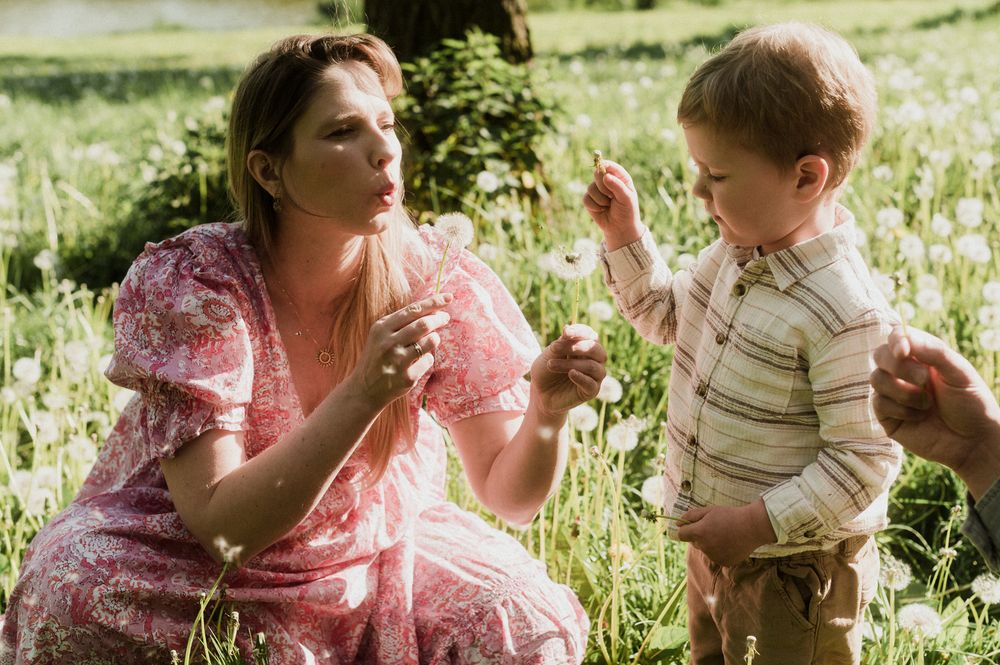 A woman in a pink floral dress kneels in a meadow, showing a flower to a young toddler boy outdoors.