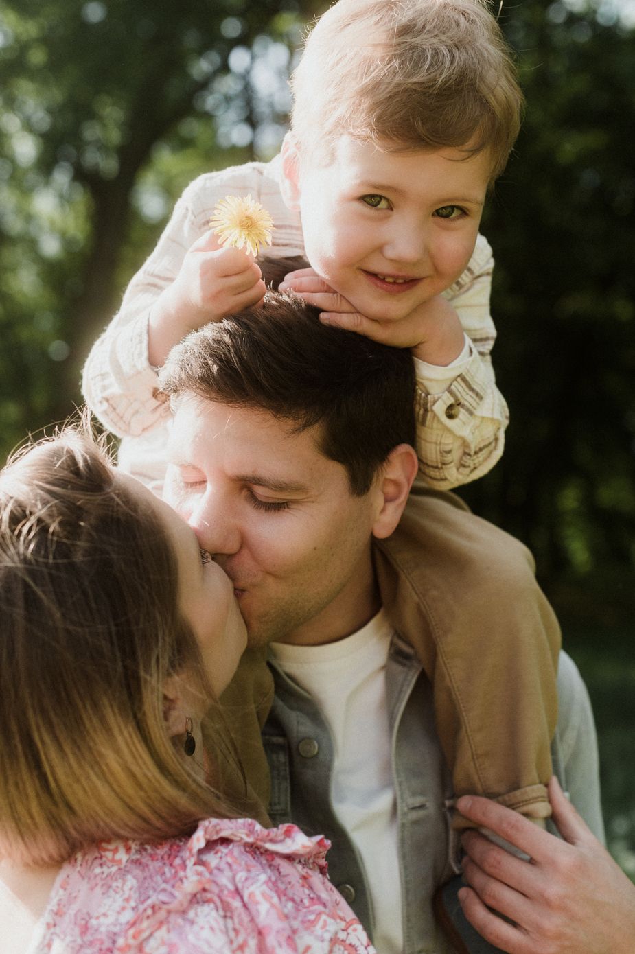 A couple shares a kiss while their toddler sits on dad's shoulders holding a flower outdoors.