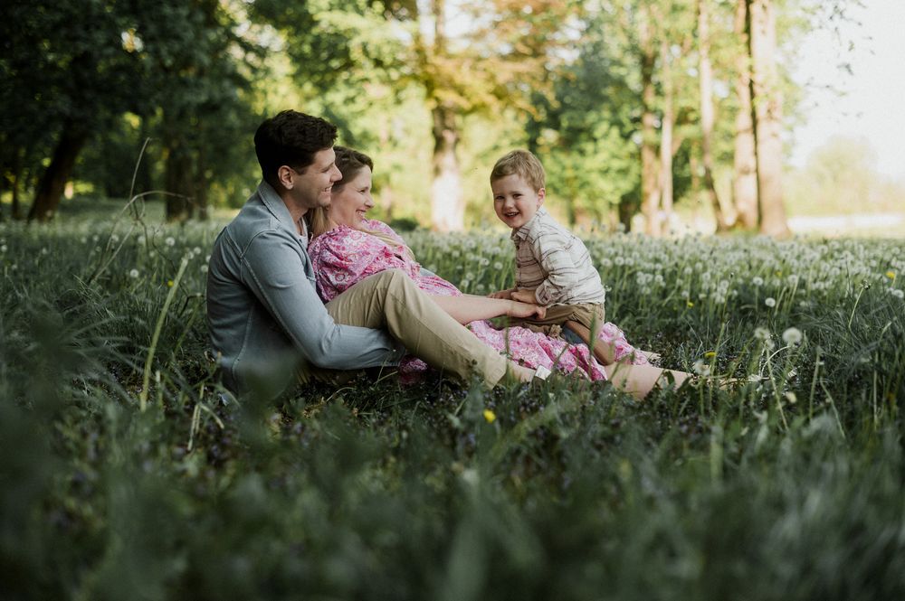 Family of three sitting together in lush green grass surrounded by trees on a sunny day.
