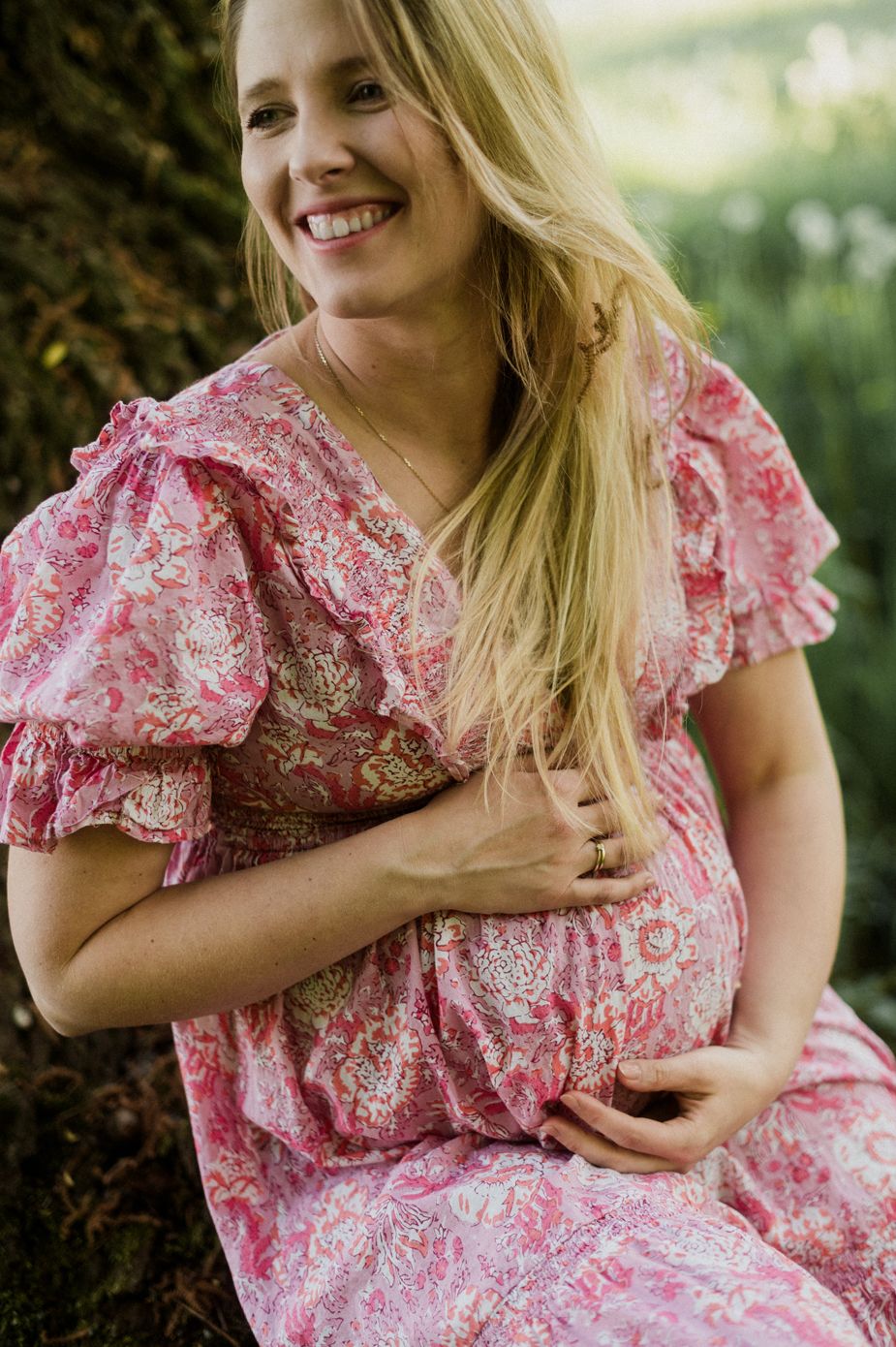 Smiling pregnant woman in pink floral dress cradling baby bump outdoors.