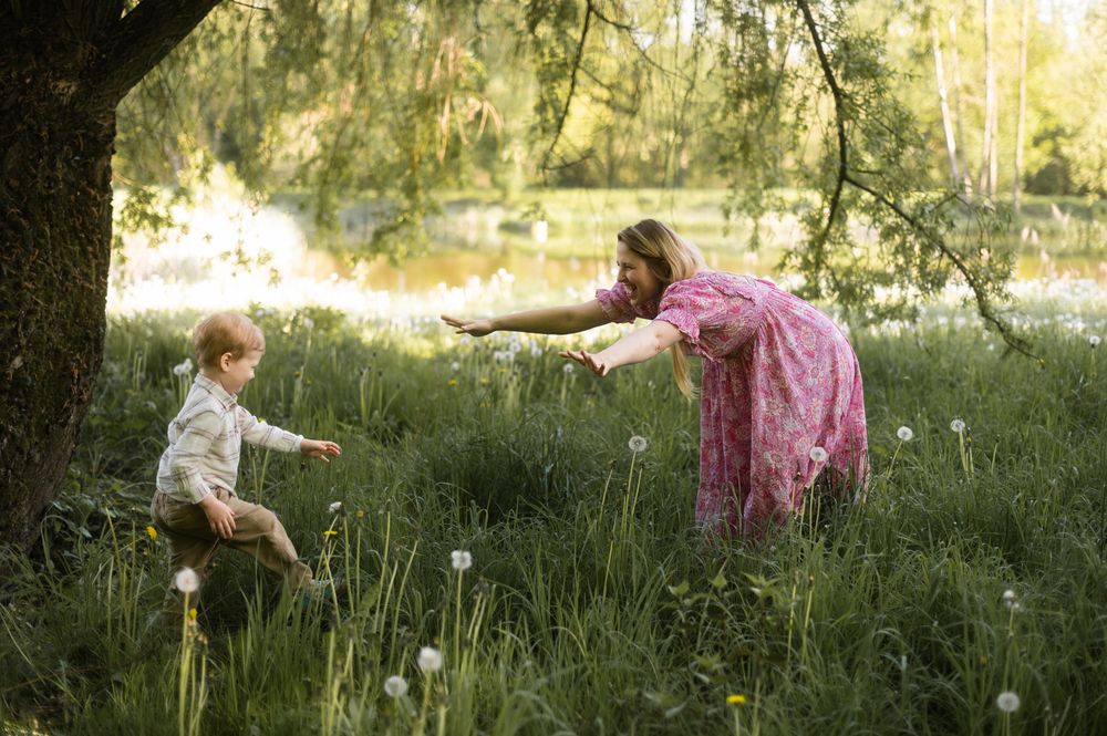 Woman in pink dress reaches out to toddler in tall grass near sunlit pond.