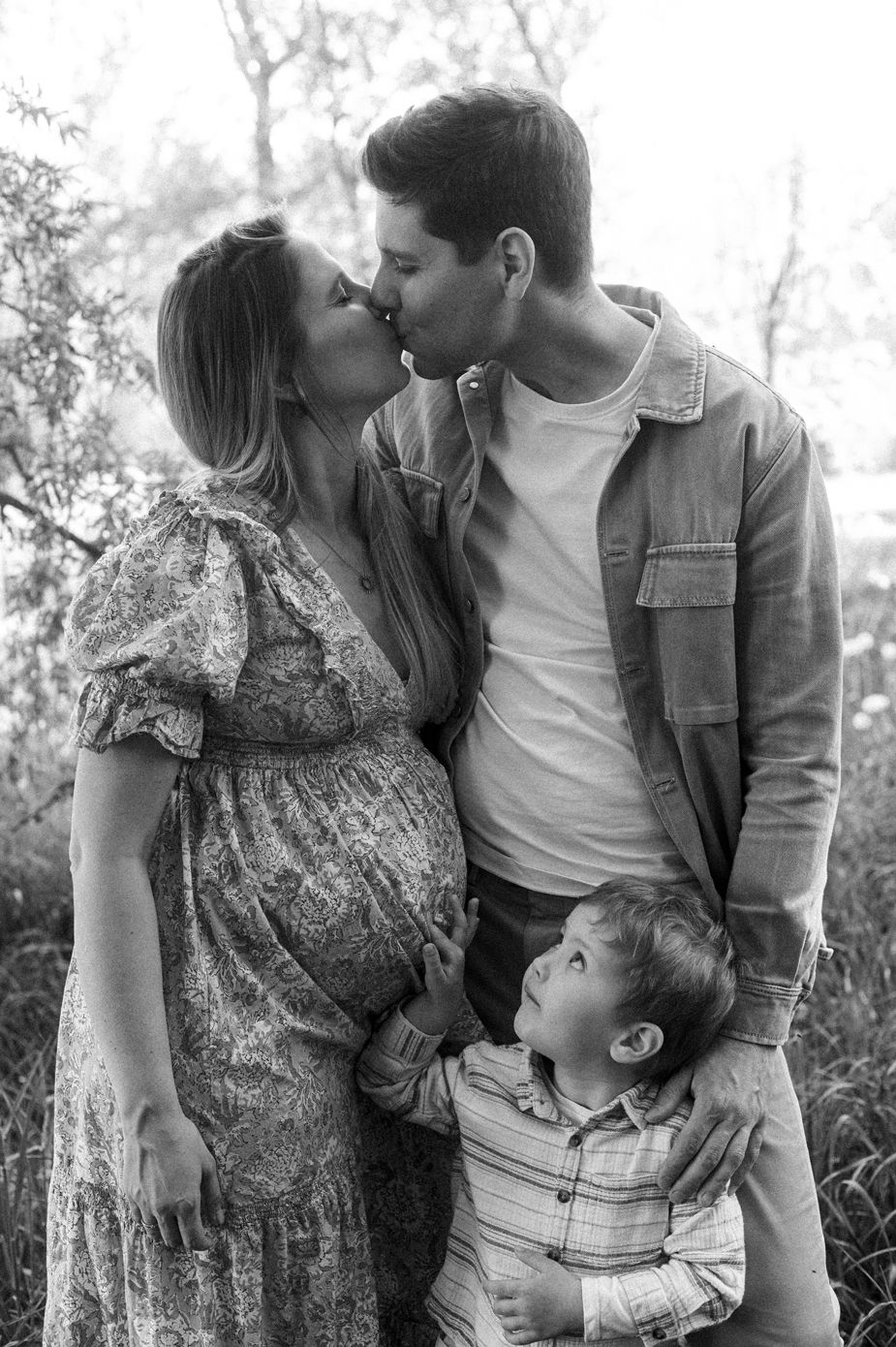 Black and white maternity photo of pregnant couple kissing outdoors with young toddler son standing between them.