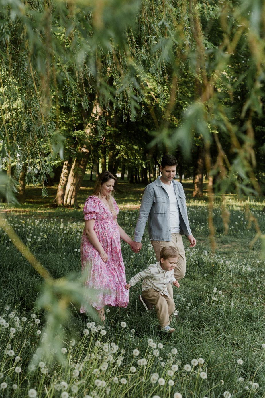 A pregnant woman in pink dress walks with husband and young son through a lush green park with dandelions.