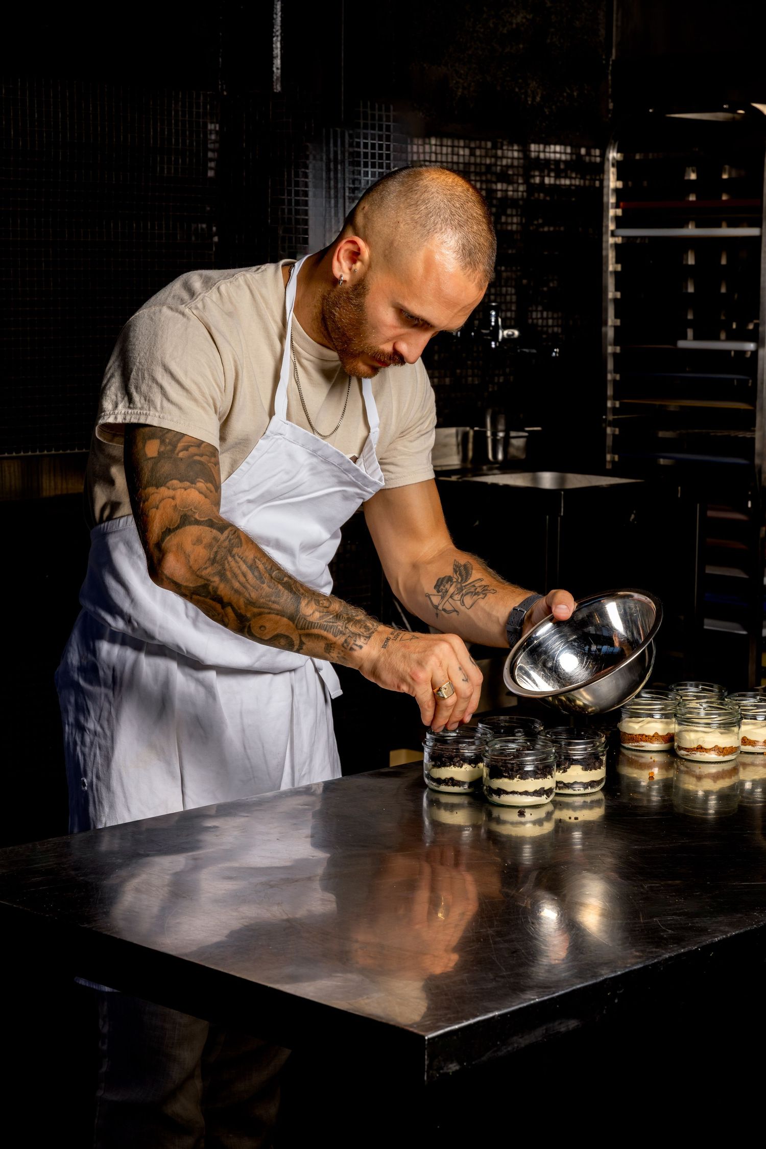 Tattooed chef in white apron carefully fills glass jars with layered desserts in a professional kitchen.
