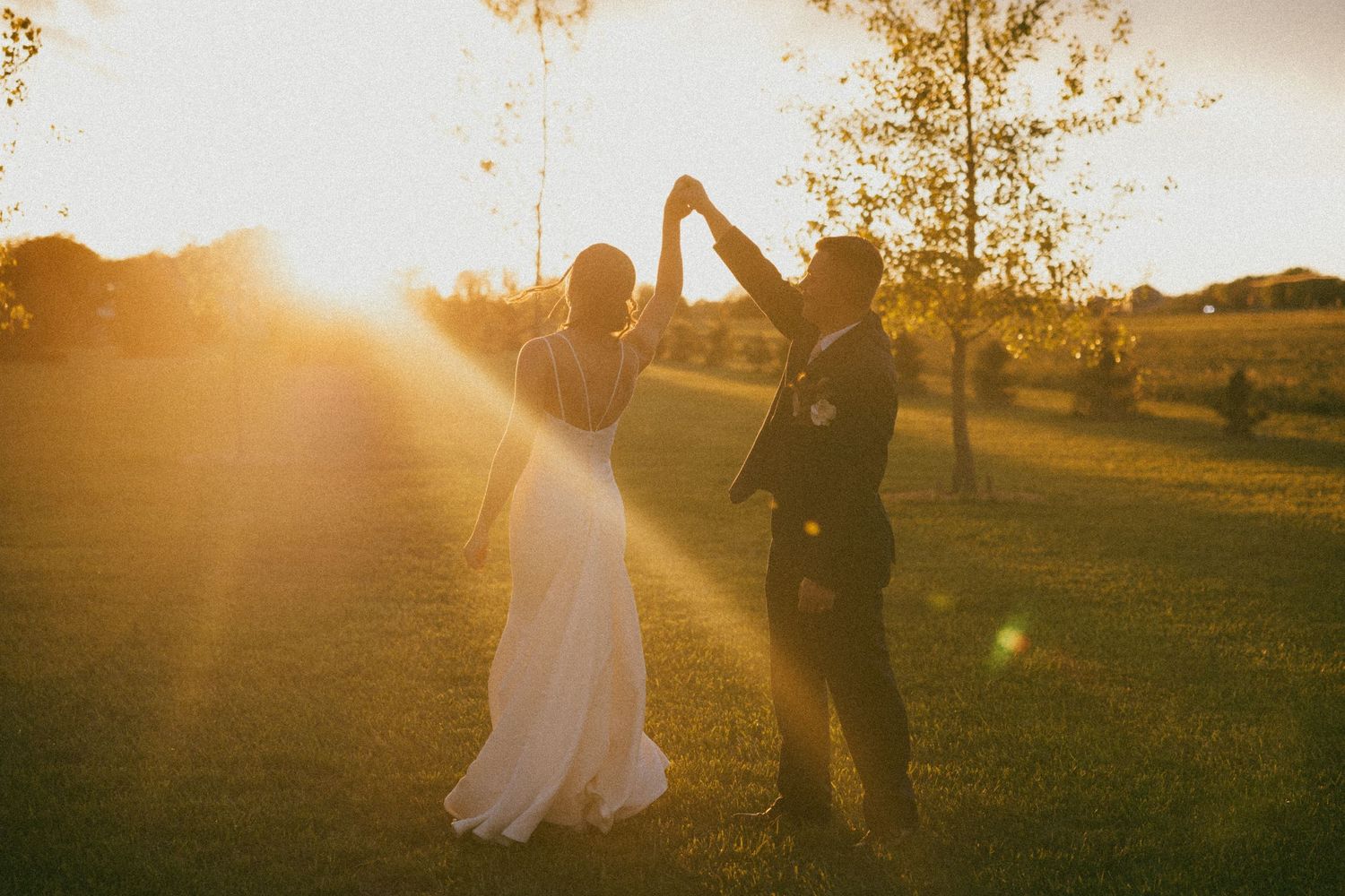 Bride and groom dancing at golden sunset in lush green field, sun flare illuminating romantic outdoor wedding celebration.