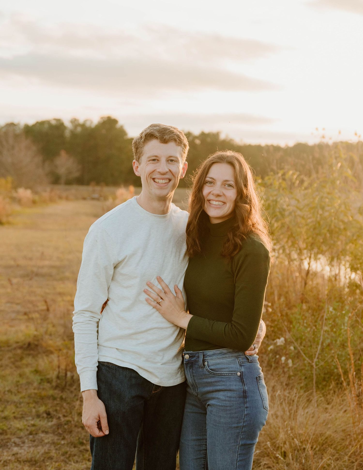 A smiling couple poses together in a golden sunset field, woman wearing green turtleneck and jeans, man in white sweater.