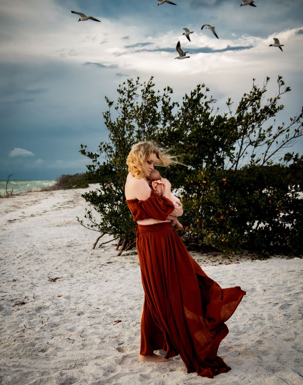 Woman in red dress cradling baby on beach with seagulls flying overhead.