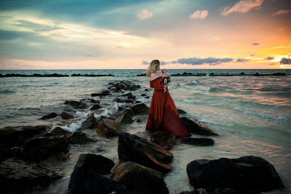 Woman in flowing red dress standing on rocks by ocean at sunset.
