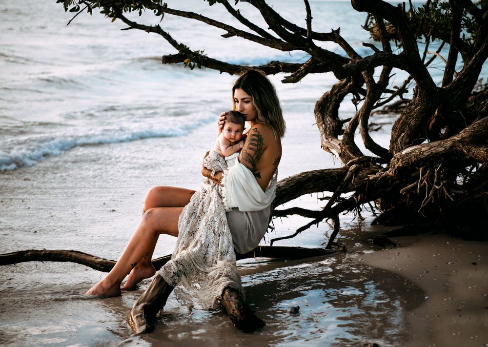 Tattooed woman sitting on driftwood at the beach holding her baby.