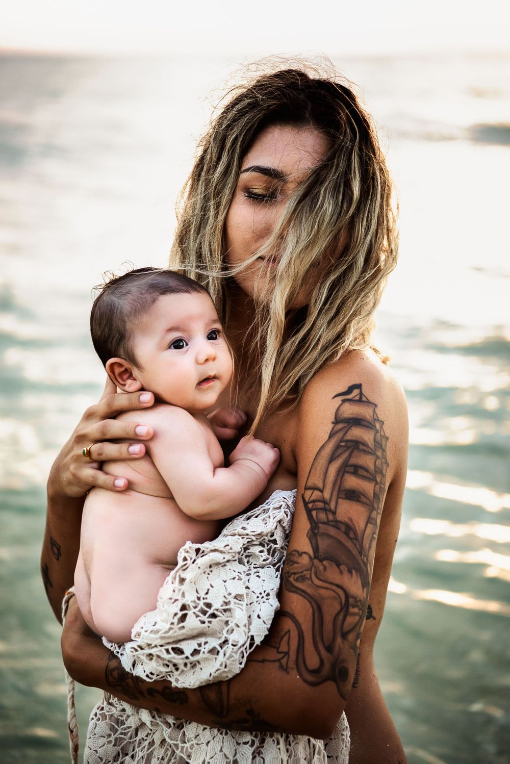 Woman with tattoos holding a newborn baby at the beach during golden hour.