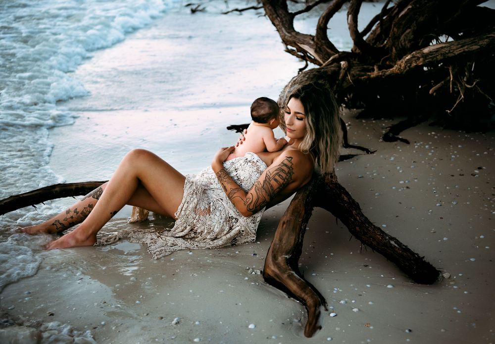 Woman with tattoos cradling a newborn baby while reclining against driftwood on a beach.