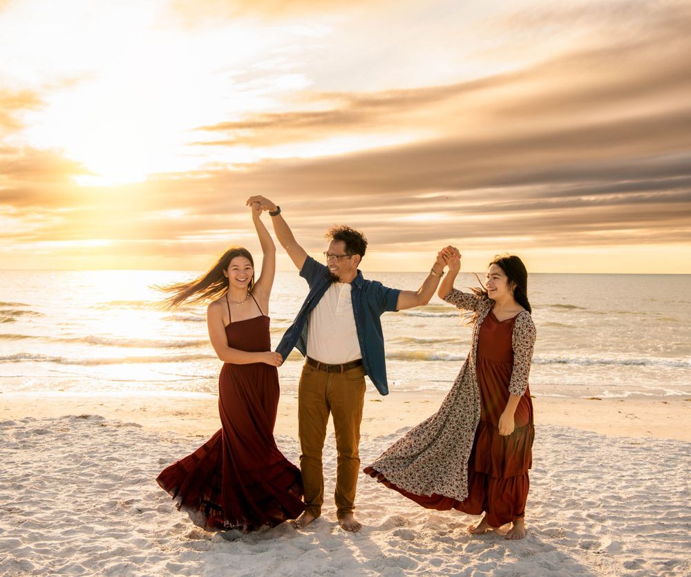 Man dancing with two women in dresses on beach at sunset.