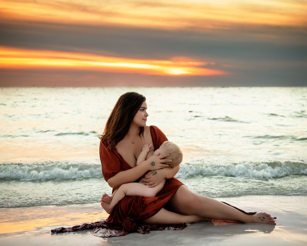 Mother sitting on beach sand nursing her baby during a vibrant sunset.