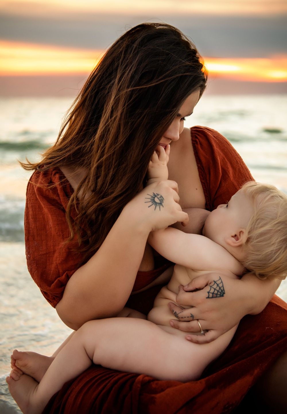 Mother in red dress breastfeeding her baby on the beach at sunset.