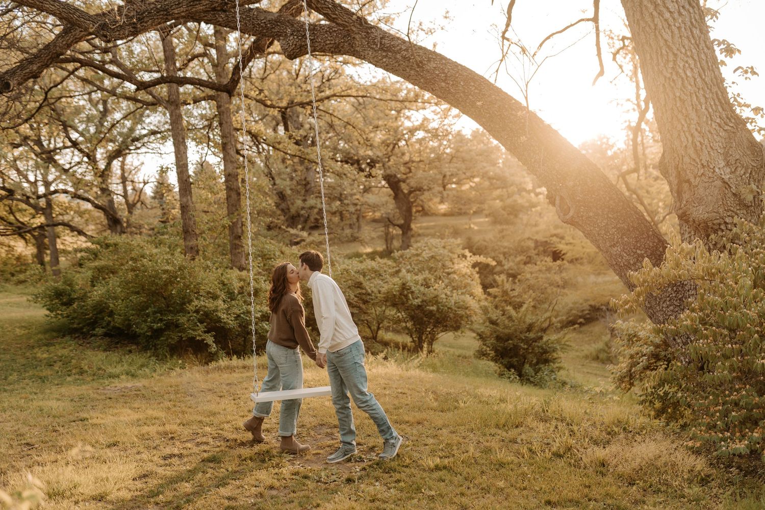 Couple holding hands near a rope swing under a large oak tree in golden sunset light in a lush green park.