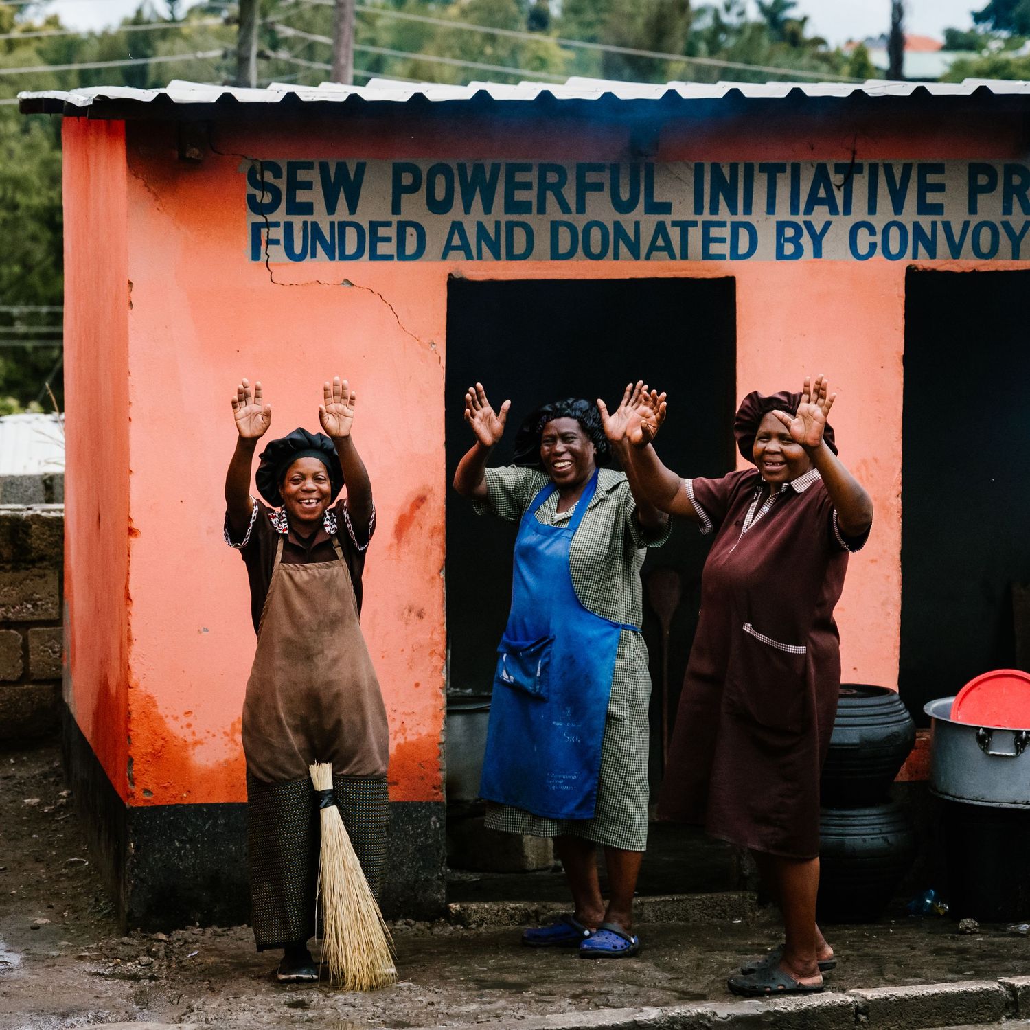 Three joyful women wave outside a colorful Sew Powerful Initiative building funded by Convoy of Hope in Africa.