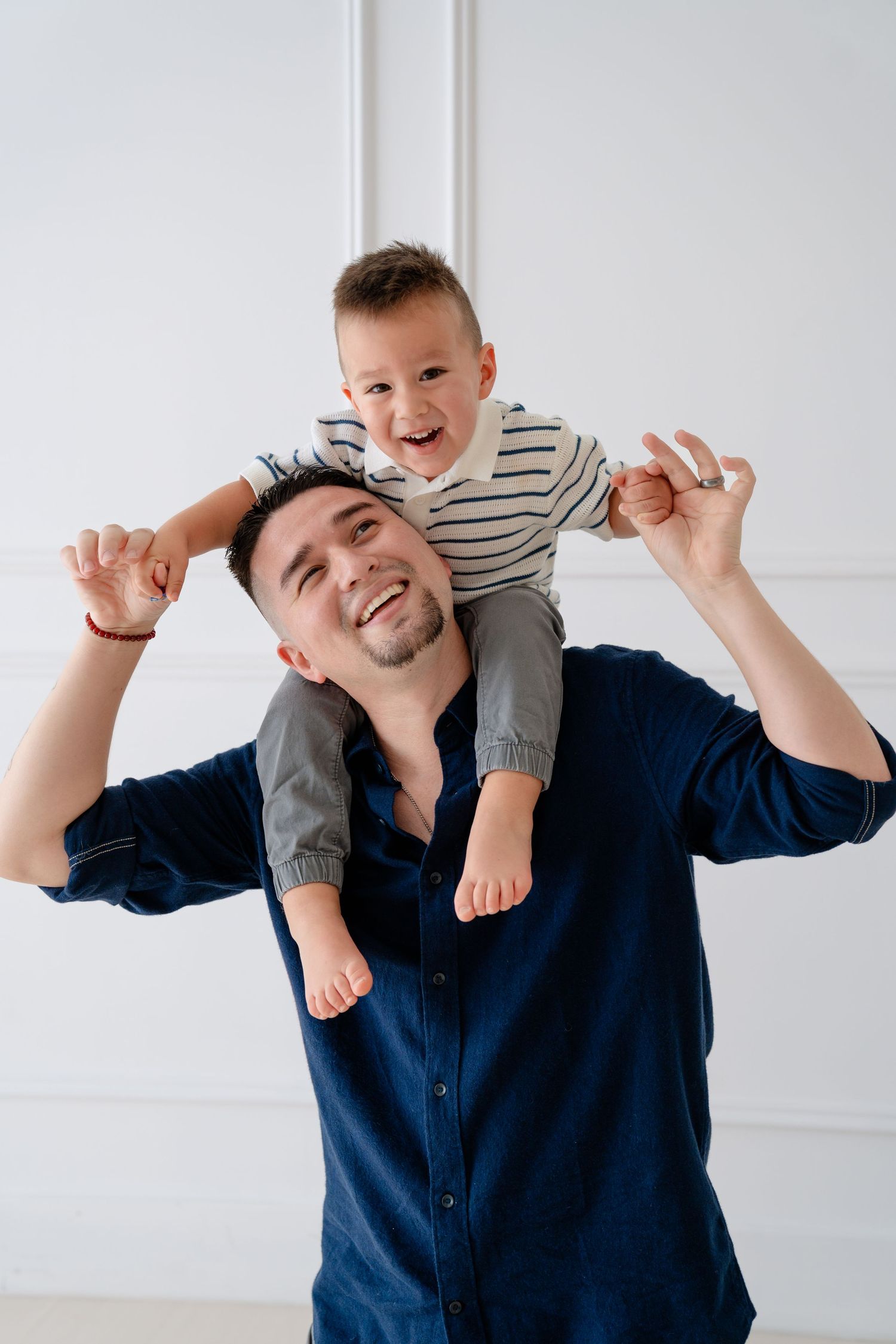 Happy father carrying laughing toddler son on shoulders in bright white room, joyful family bonding moment.