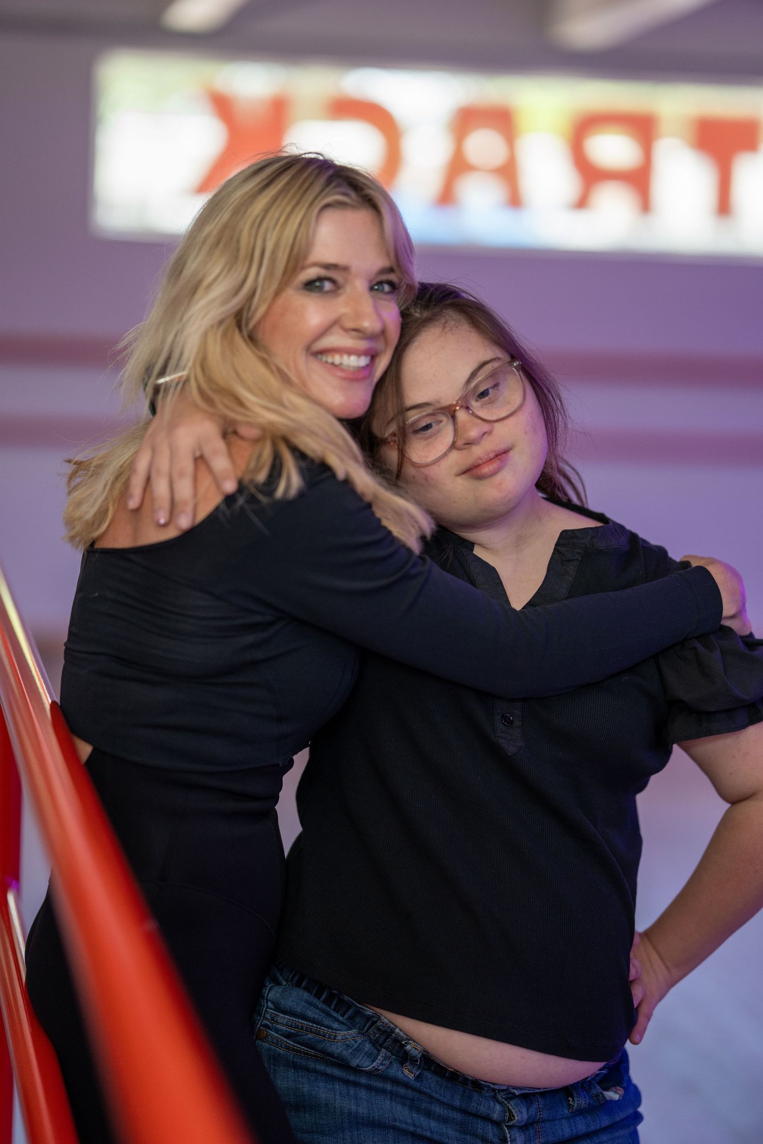 Blonde woman in black top gesturing while speaking under purple lighting in a dance studio.