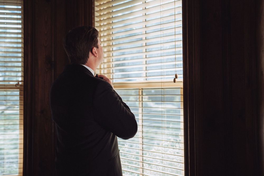 Man in suit standing by window with blinds, looking outside in a contemplative pose.