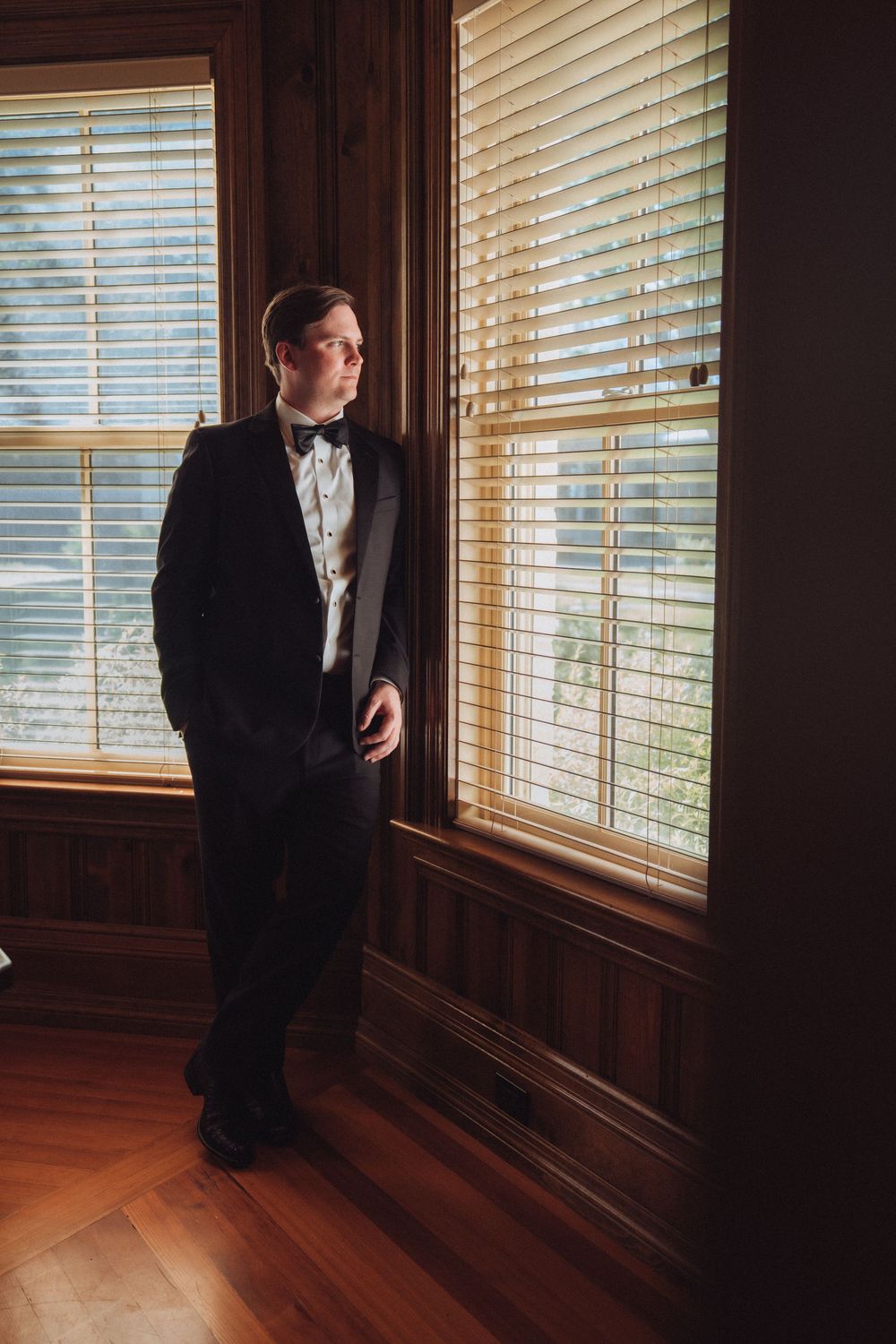 Groom in black tuxedo and bow tie standing by wooden window with blinds in elegant room.