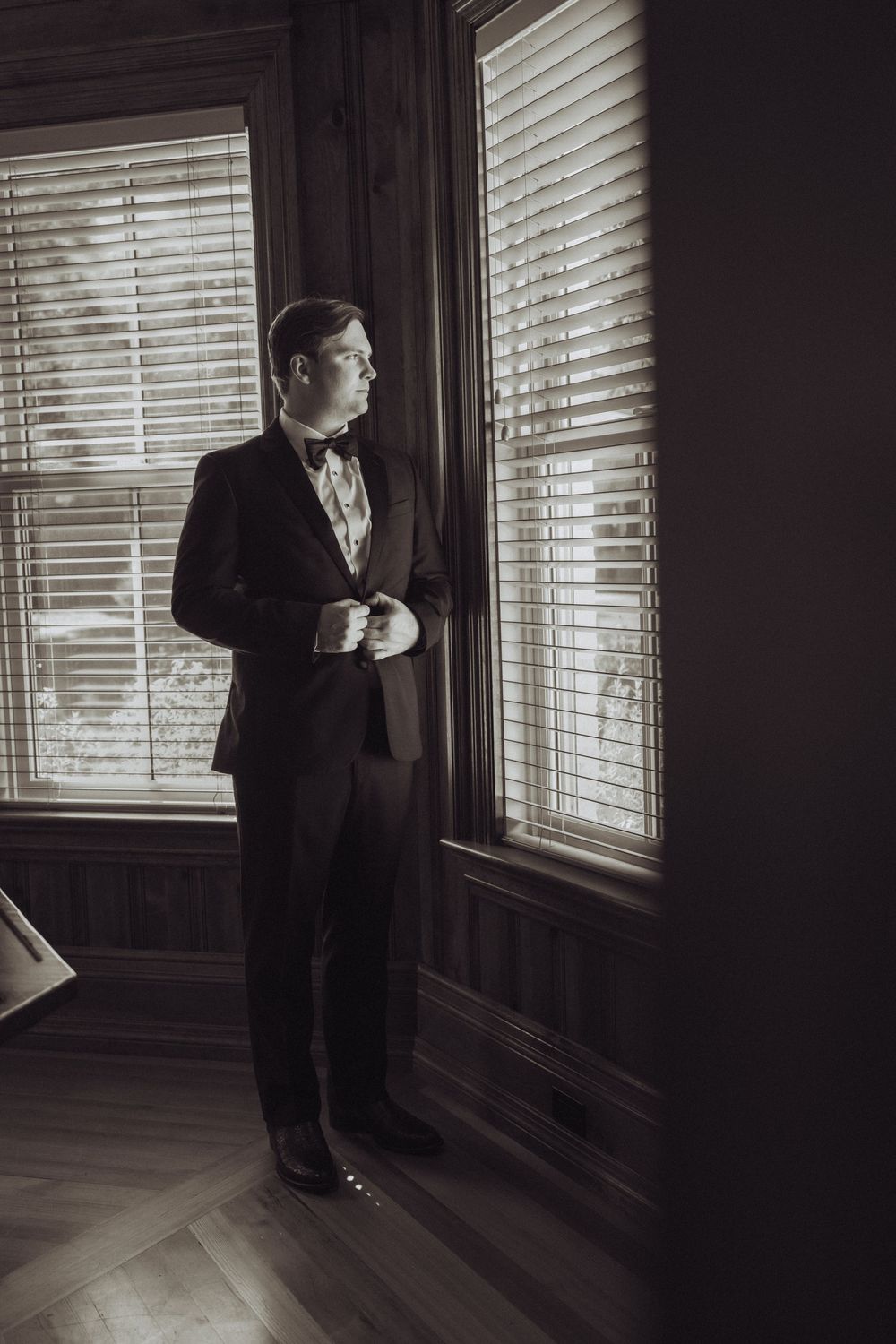 Groom in black tuxedo and bow tie standing by window with blinds in elegant dark wood room.