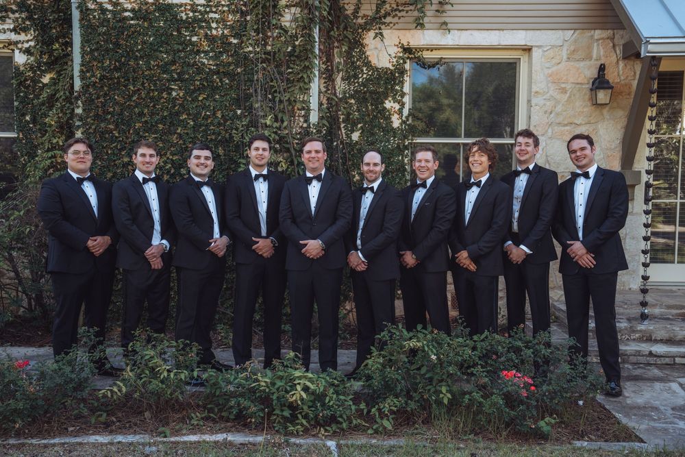 A group of ten groomsmen in black tuxedos and bow ties posing together outside a stone building.